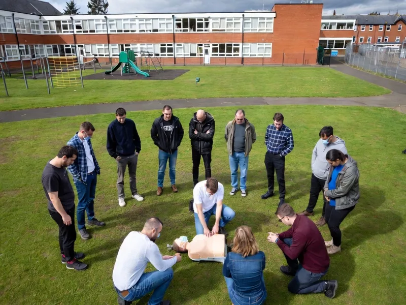 Outdoor first aid training session at a school, with learners gathered around a CPR demonstration on the grass